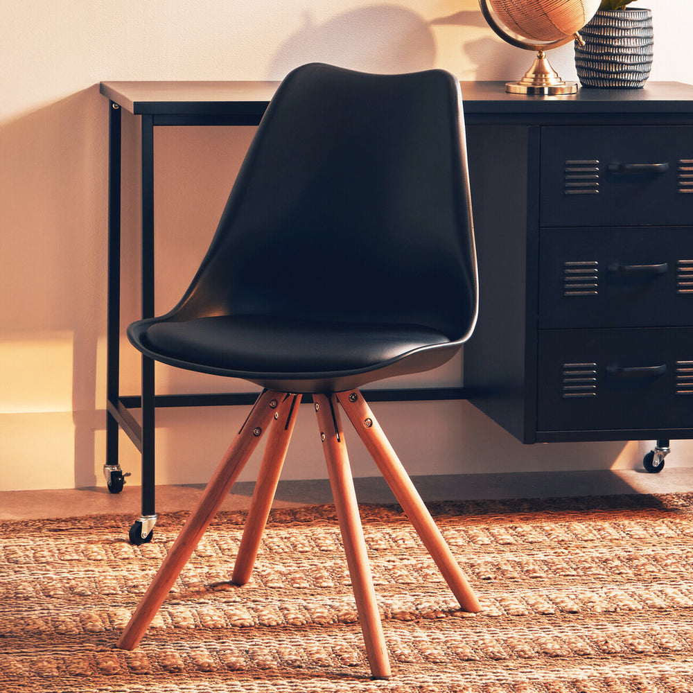 Black padded seat dining chair with a sleek, curved backrest and wooden legs, positioned on a textured rug in a well-lit room with a side table and decorative items in the background