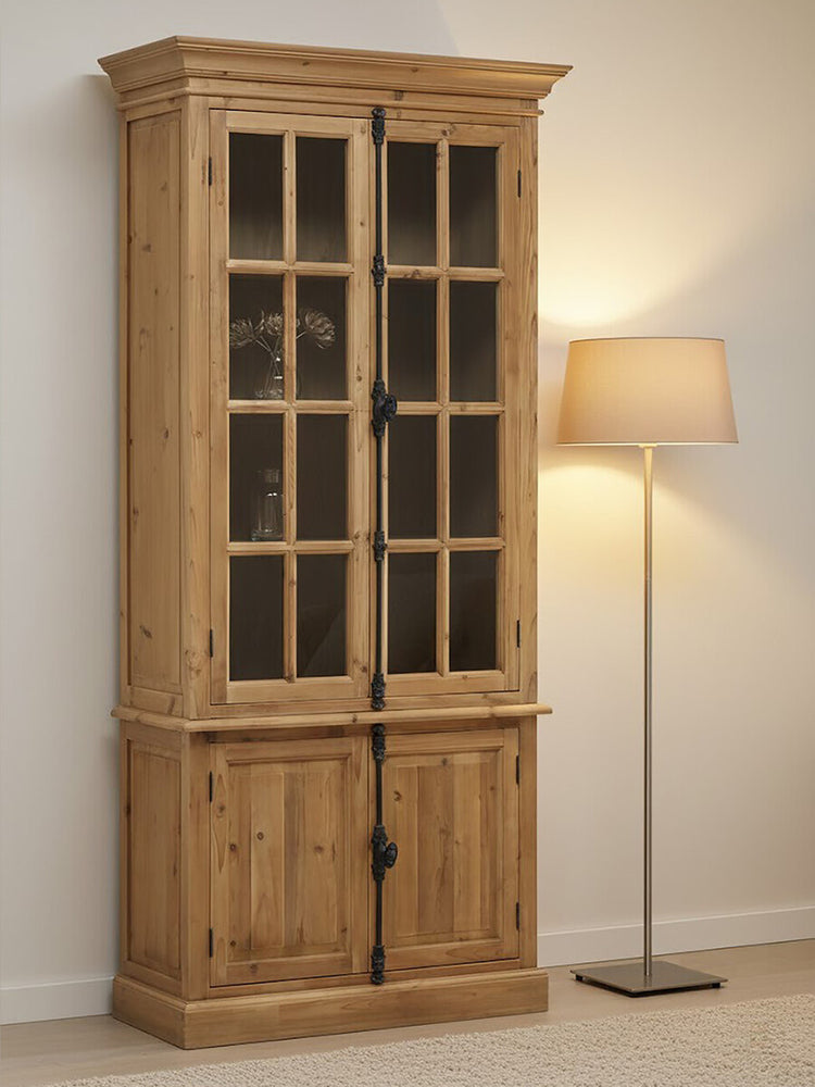 Tall cabinet made of pine wood featuring four doors, two with glass panels and two solid wood doors at the bottom. The cabinet has a natural wood finish, showcasing the grain, and is positioned next to a floor lamp with a soft, warm light. The background is a neutral wall, enhancing the cabinet's rustic charm