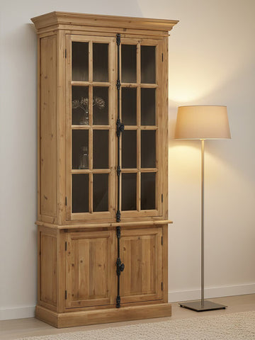 Tall cabinet made of pine wood featuring four doors, two with glass panels and two solid wood doors at the bottom. The cabinet has a natural wood finish, showcasing the grain, and is positioned next to a floor lamp with a soft, warm light. The background is a neutral wall, enhancing the cabinet's rustic charm