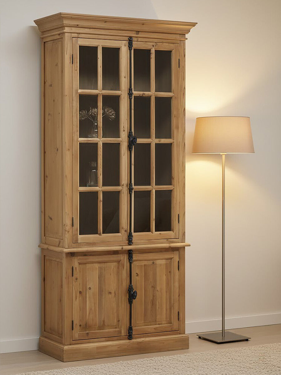 Tall cabinet made of pine wood featuring four doors, two with glass panels and two solid wood doors at the bottom. The cabinet has a natural wood finish, showcasing the grain, and is positioned next to a floor lamp with a soft, warm light. The background is a neutral wall, enhancing the cabinet's rustic charm