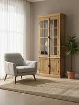 A tall cabinet made of pine wood with four glass doors, showcasing a natural wood finish. The cabinet is positioned next to a light gray armchair with wooden legs, on a beige area rug. A potted plant is visible in the background, and sunlight filters through sheer curtains, creating a warm and inviting atmosphere
