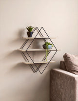 Three-tier rhombus wall shelf mounted on a light-colored wall, featuring two small potted plants on the top shelf and a minimalist design with a geometric metal frame. A corner of a beige sofa is visible in the foreground