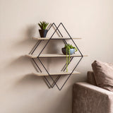 Three-tier wall shelf with a rhombus design, featuring light wooden shelves supported by a black metal frame. Two small potted plants are placed on the top and middle shelves. A beige sofa is partially visible on the right side against a light-colored wall