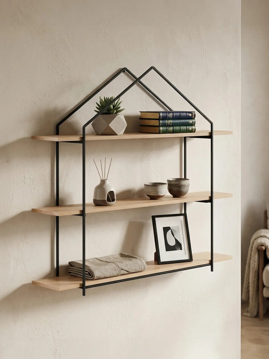 Three-tier wooden wall shelves with a black metal frame shaped like a house. The shelves are made of light-colored wood and display decorative items, including a small plant, books, and a framed photo. The background features a neutral wall, enhancing the modern aesthetic of the shelves