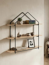 Three-tier wooden wall shelves with a black metal frame shaped like a house. The shelves are made of light-colored wood and display decorative items, including a small plant, books, and a framed photo. The background features a neutral wall, enhancing the modern aesthetic of the shelves