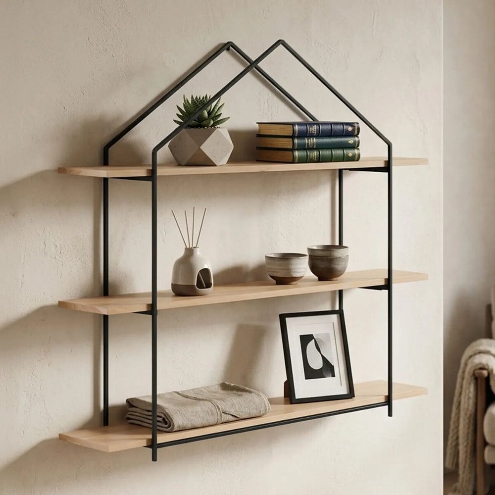Three-tier wooden wall shelves with a black metal frame designed in a house shape, featuring natural wood shelves displaying a small potted plant, stacked books, and a framed picture, mounted against a light-colored wall