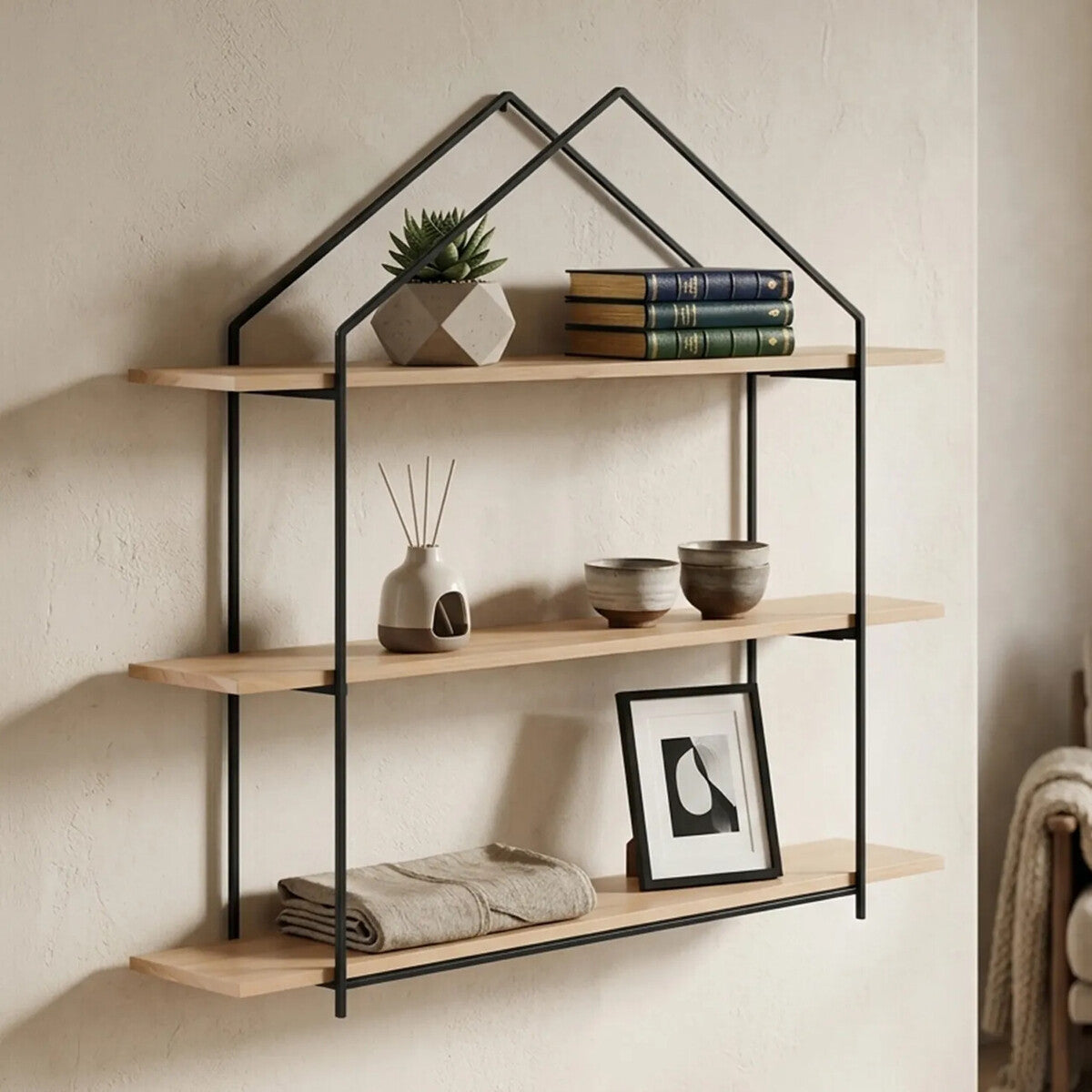 Three-tier wooden wall shelves with a black metal frame designed in a house shape, featuring natural wood shelves displaying a small potted plant, stacked books, and a framed picture, mounted against a light-colored wall