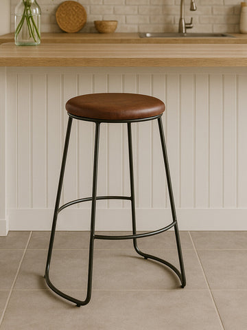 Brixton faux leather bar stool featuring a round brown seat and sleek black metal legs, positioned on a light-colored tiled floor with a wooden countertop in the background