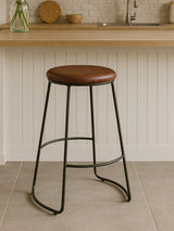 Brixton faux leather bar stool featuring a round brown seat and sleek black metal legs, positioned on a light-colored tiled floor with a wooden countertop in the background