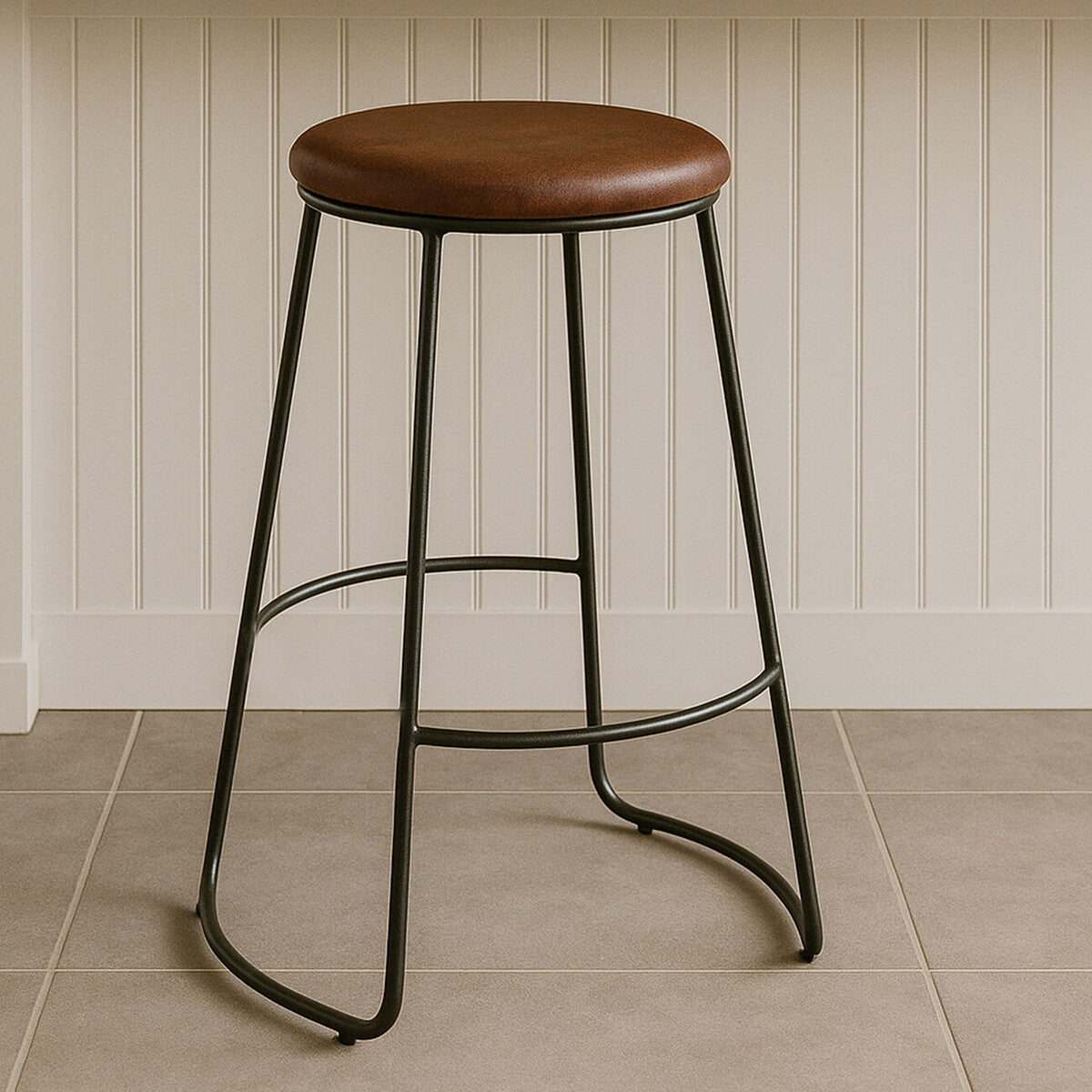 Brixton faux leather bar stool featuring a round brown seat and sleek black metal legs, positioned on a light-colored tiled floor against a white panelled wall