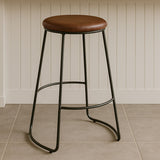 Brixton faux leather bar stool featuring a round brown seat and sleek black metal legs, positioned on a light-colored tiled floor against a white panelled wall