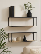 Two modern cuboid wall shelves in a black metal frame, mounted on a light-colored wall. The top shelf holds a black cylindrical vase and a small potted plant, while the bottom shelf displays a white decorative orb, a black vase, and a beige decorative piece. A green plant is partially visible in the foreground