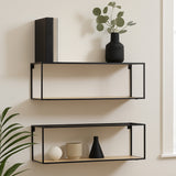 Two cuboid wall shelves in a modern design, featuring a black metal frame with wooden shelves. The top shelf displays a black book and a black vase with greenery, while the bottom shelf holds a white decorative orb and a black triangular object. The shelves are mounted against a light-colored wall, with a green plant partially visible in the foreground
