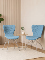 Two Stockholm blue dining chairs with a quilted design are positioned around a small round table. A potted plant sits on the table, and the background features light-colored walls with subtle texture. A soft area rug is visible beneath the chairs