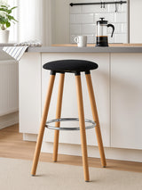 Black round bar stool with a cushioned seat and wooden legs, featuring a chrome footrest, positioned next to a kitchen counter with a coffee maker and a white mug in the background