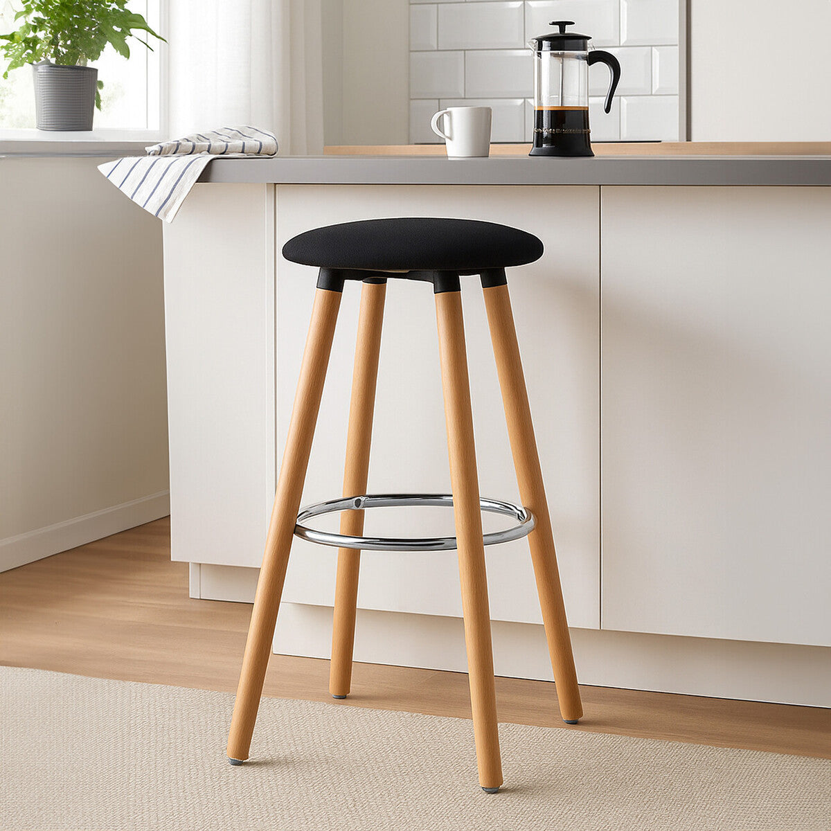 Black round bar stool with a cushioned seat and wooden legs, featuring a chrome footrest, positioned next to a kitchen counter with a coffee maker and a plant in the background. The floor is light-colored and the walls are neutral
