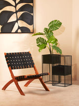 A black woven chair with a wooden frame is positioned next to a modern black wire side table, adorned with a leafy green plant. The background features a light-colored wall and a large piece of wall art with abstract designs in black and beige tones