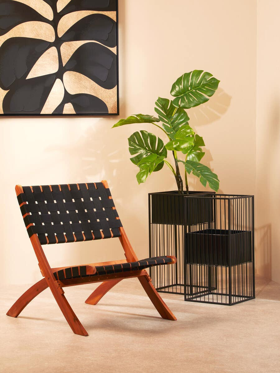 A black woven chair with a wooden frame is positioned next to a modern black wire side table, adorned with a leafy green plant. The background features a light-colored wall and a large piece of wall art with abstract designs in black and beige tones