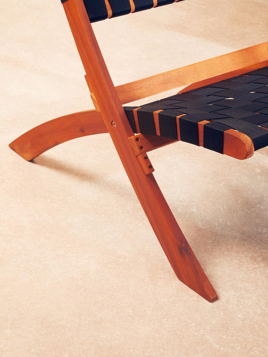 A close-up view of the Emilio Black Woven Chair, showcasing its black woven seat and backrest, supported by a light wooden frame with a curved design. The chair is positioned on a soft, neutral-colored floor
