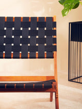 Black woven chair featuring a slatted backrest and seat, constructed with a wooden frame. The woven design includes contrasting lighter accents, set against a neutral background with a decorative plant and a modern metal side table nearby