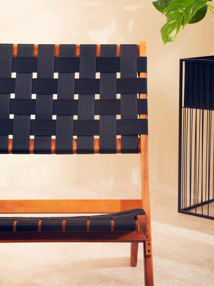 Black woven chair featuring a slatted backrest and seat, constructed with a wooden frame. The woven design includes contrasting lighter accents, set against a neutral background with a decorative plant and a modern metal side table nearby