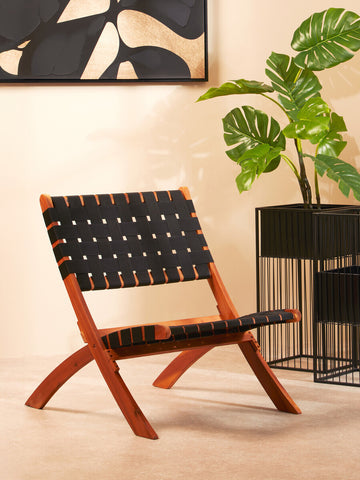 A black woven chair with a wooden frame, featuring a minimalist design and crossed legs, positioned in a stylish interior setting with a large abstract wall art piece and a potted plant nearby