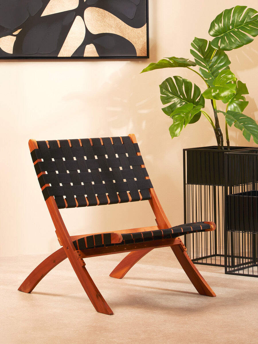 A black woven chair with a wooden frame, featuring a minimalist design and crossed legs, positioned in a stylish interior setting with a large abstract wall art piece and a potted plant nearby
