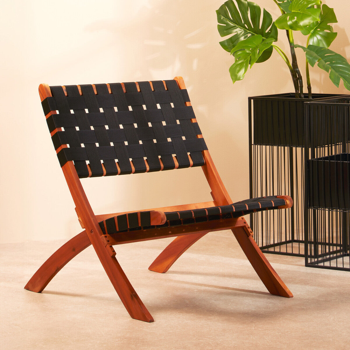 A black woven chair with a wooden frame featuring a cross-legged design, set against a light-colored wall. The chair has a modern aesthetic with a woven seat and backrest, complemented by a nearby potted plant and a metal side table