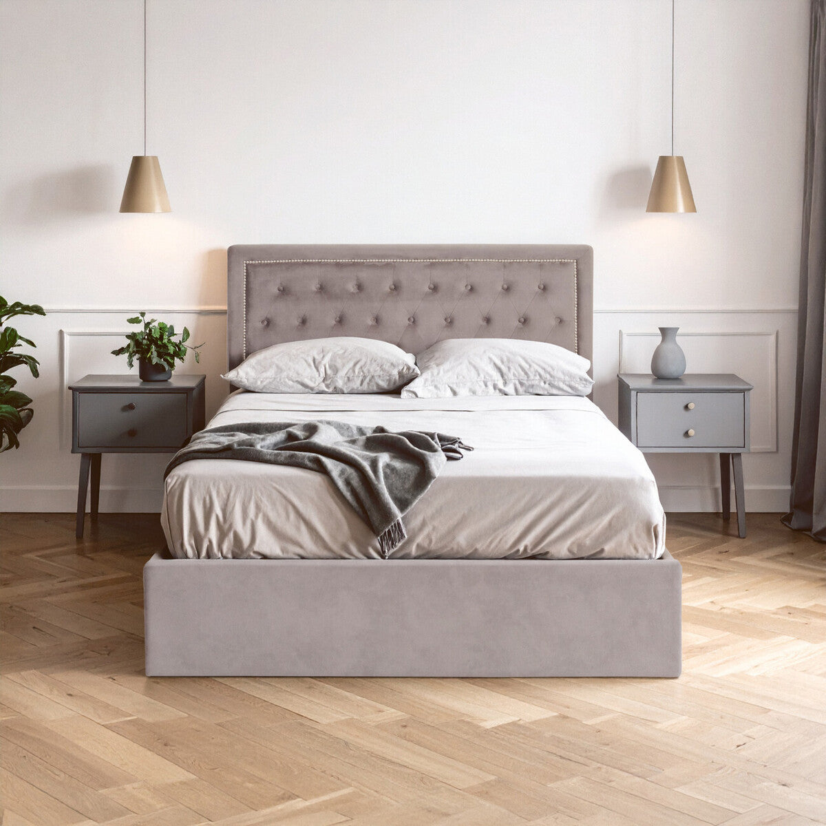 Grey velvet double ottoman bed with a tufted headboard, neatly made with white bedding and a dark throw blanket, positioned between two modern bedside tables with lamps, set against a light-colored wall and wooden floor