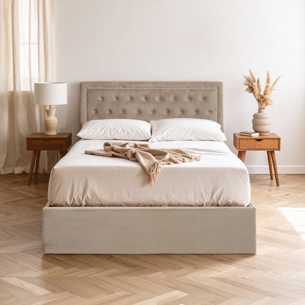 Mink velvet double bed with a tufted headboard, white bedding, and decorative throw draped across the top. The bed is positioned on a light wooden floor, accompanied by two wooden nightstands with a lamp and a vase of dried flowers. Soft natural light filters through sheer curtains in the background