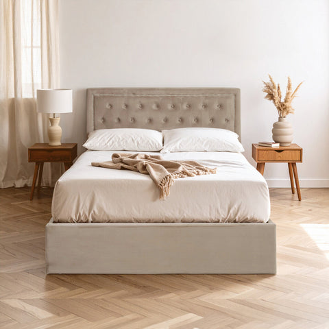 Mink velvet double bed with a tufted headboard, white bedding, and decorative throw draped across the top. The bed is positioned on a light wooden floor, accompanied by two wooden nightstands with a lamp and a vase of dried flowers. Soft natural light filters through sheer curtains in the background