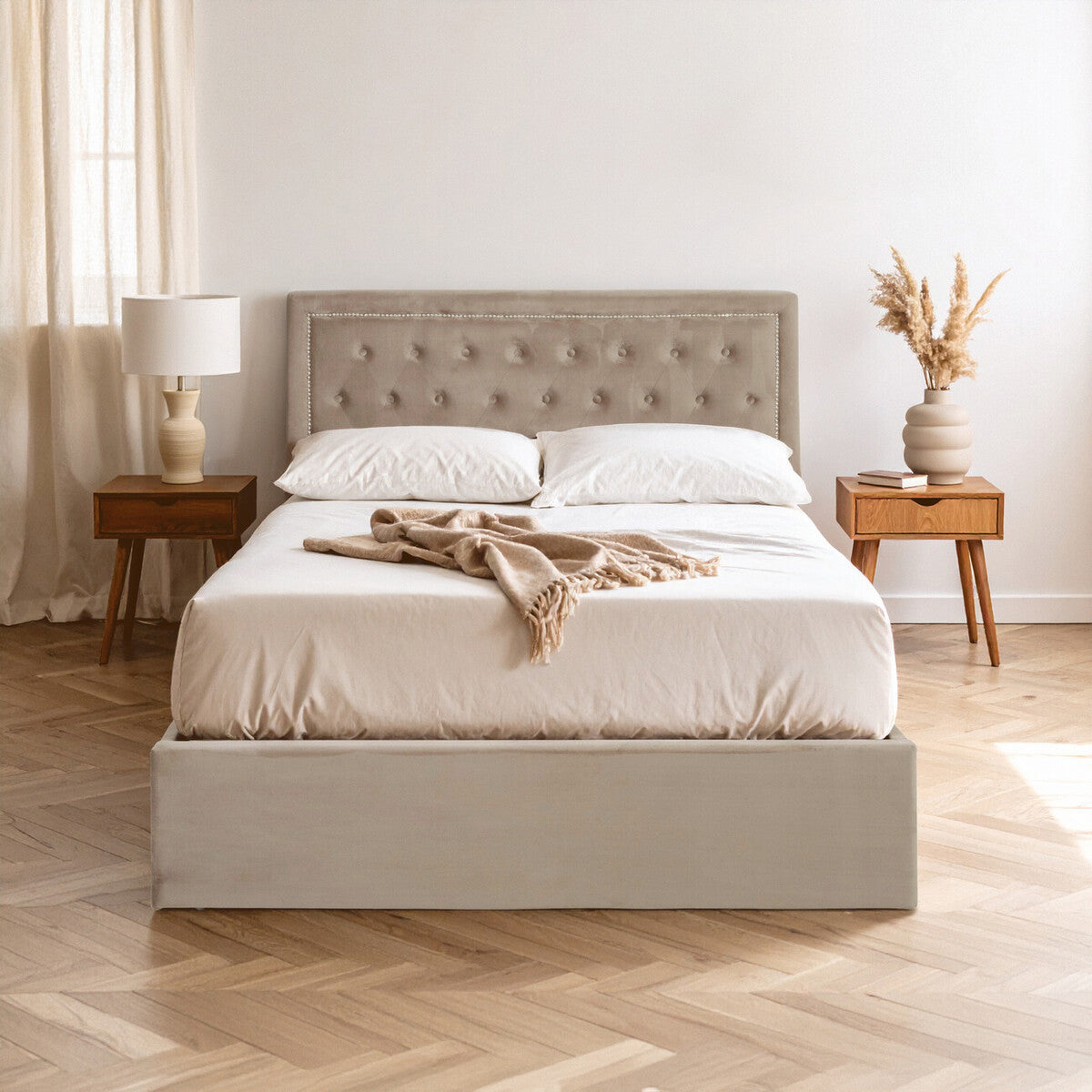 Mink velvet double bed with a tufted headboard, white bedding, and decorative throw draped across the top. The bed is positioned on a light wooden floor, accompanied by two wooden nightstands with a lamp and a vase of dried flowers. Soft natural light filters through sheer curtains in the background
