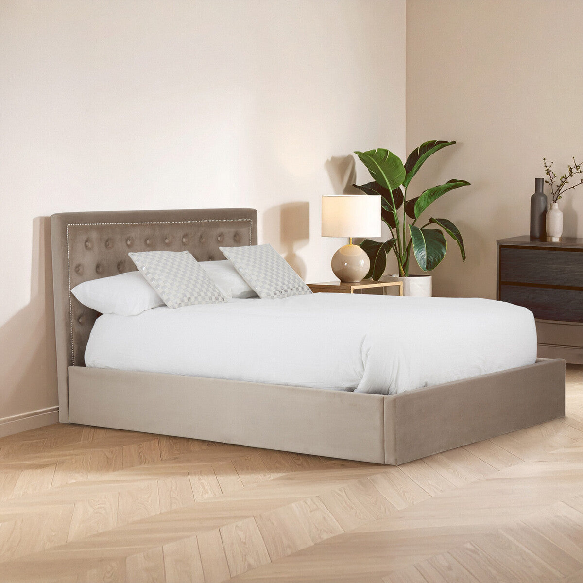 Mink velvet king bed with a tufted headboard, featuring a white comforter and two decorative pillows, set against a light-colored wall. A bedside table with a lamp and a potted plant is visible in the background, along with a wooden floor
