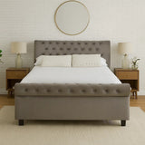 Grey upholstered king bed with a tufted headboard, featuring a padded ottoman base. The bed is positioned on a light-colored rug, flanked by two wooden nightstands with lamps. A round mirror is mounted on the wall above the bed