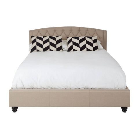 Beige king bed with a tufted headboard, featuring a white mattress and decorative black and white chevron-patterned pillows. The bed is supported by dark wooden legs