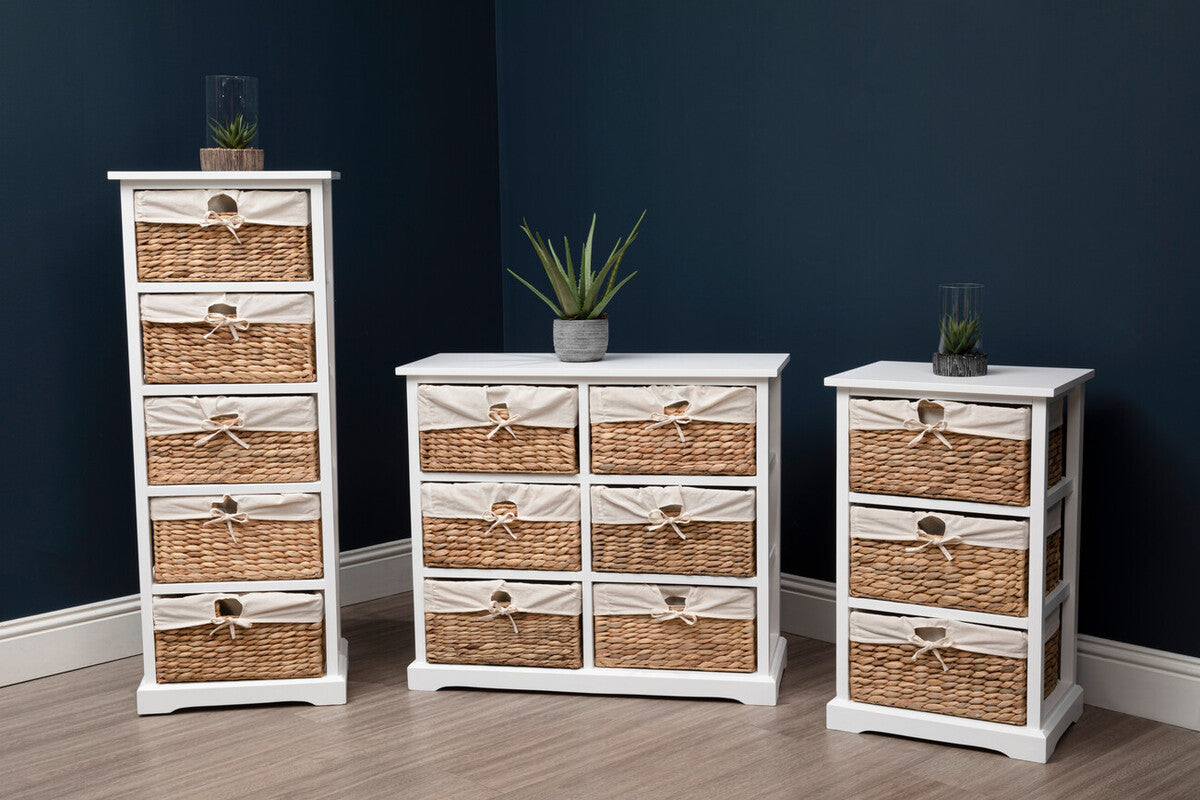 A white wooden chest of drawers featuring three seagrass baskets, positioned in a modern room with a dark blue wall. The chest is flanked by two additional storage units, all showcasing a combination of white frames and natural seagrass. A small potted plant sits atop the chest, adding a touch of greenery to the scene