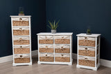 A white wooden chest of drawers featuring three seagrass baskets, positioned in a modern room with a dark blue wall. The chest is flanked by two additional storage units, all showcasing a combination of white frames and natural seagrass. A small potted plant sits atop the chest, adding a touch of greenery to the scene