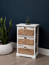 A white wooden chest with three seagrass drawers, featuring natural woven textures and small metal handles, positioned next to a large white planter with a green plant against a dark blue wall