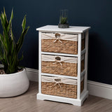 A white wooden chest with three woven seagrass drawers, featuring decorative handles, positioned next to a large potted plant on a light-colored floor against a dark blue wall