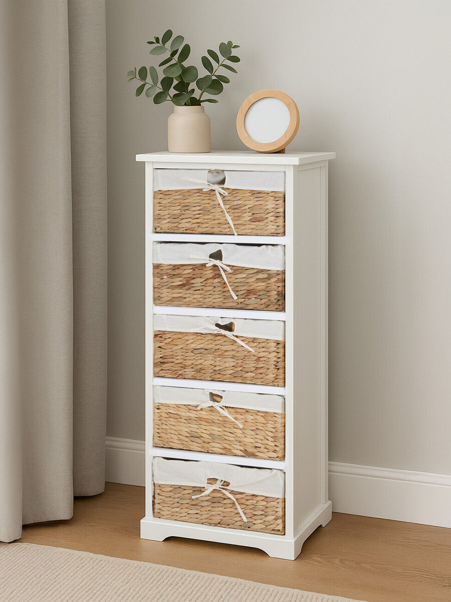 A tall, white wooden drawer chest with five woven seagrass drawers, featuring silver handles. The chest is placed against a light-colored wall, accompanied by a small potted plant and a round clock on top. A soft curtain and wooden floor are visible in the background