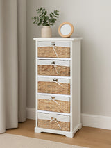 A tall, white wooden drawer chest with five woven seagrass drawers, featuring silver handles. The chest is placed against a light-colored wall, accompanied by a small potted plant and a round clock on top. A soft curtain and wooden floor are visible in the background