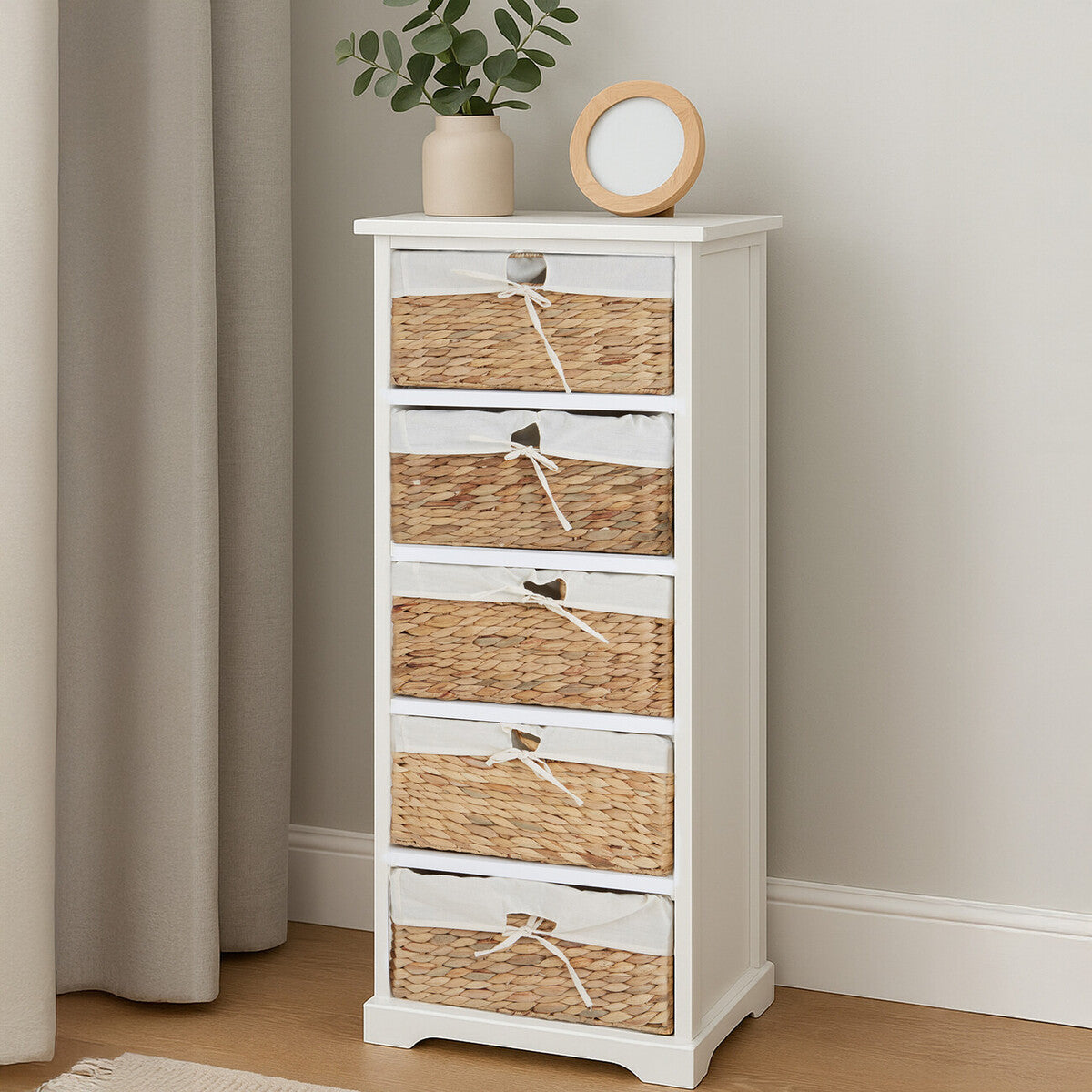 A tall, white wooden chest with five woven seagrass drawers, featuring a simple design and a small round clock and a potted plant on top, set against a light-colored wall and wooden floor