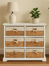 A white wooden chest of drawers featuring six seagrass-woven drawers with natural fiber handles, placed against a light-colored wall. A small potted plant and a lamp are positioned on top of the chest, adding a decorative touch to the scene