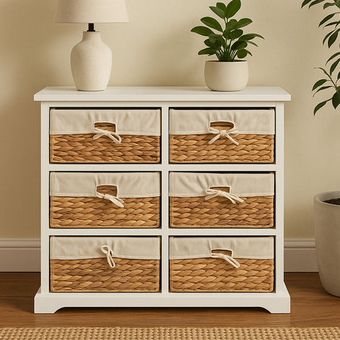 A white wooden chest of drawers featuring six seagrass-woven drawers with natural fiber accents, positioned against a light-colored wall. A small potted plant and a lamp are placed on top of the chest, adding a cozy touch to the scene