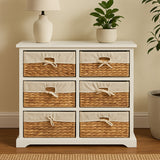 A white wooden chest of drawers featuring six seagrass-woven drawers with natural fiber accents, positioned against a light-colored wall. A small potted plant and a lamp are placed on top of the chest, adding a cozy touch to the scene