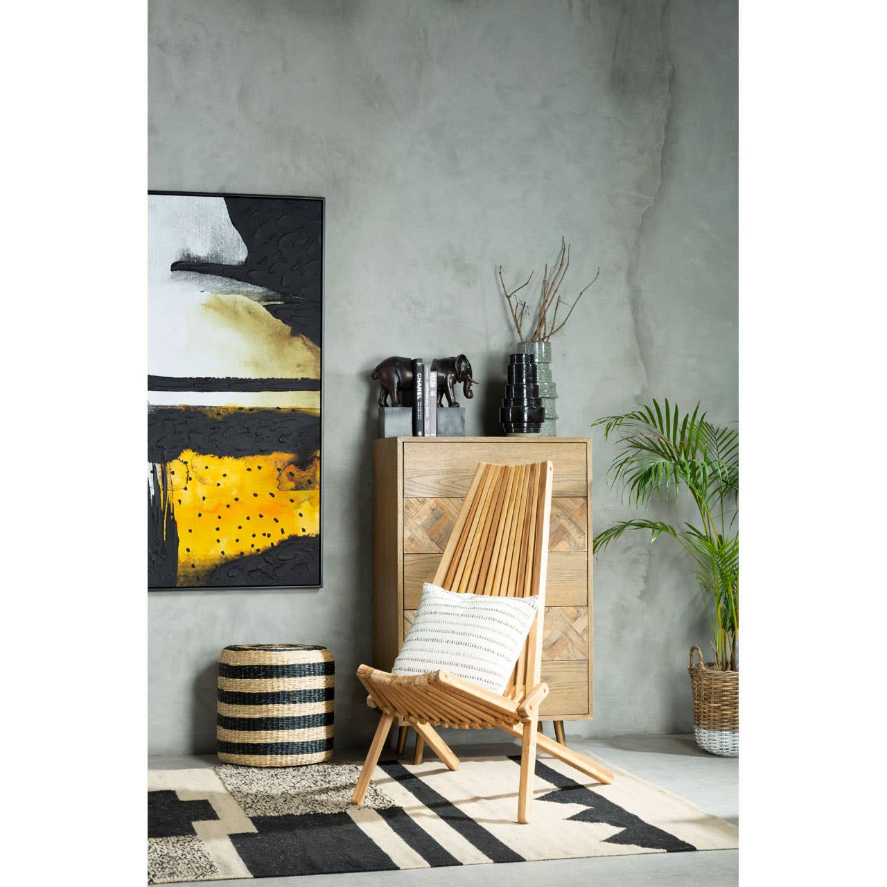 A seagrass pouffe featuring black stripes, placed on a patterned rug in a modern living space. The background includes a wooden cabinet, a stylish chair, and abstract wall art with yellow and black tones. A potted plant adds a touch of greenery to the scene