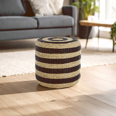 Round pouffe made of seagrass featuring alternating black and natural-colored stripes, placed on a light wooden floor in a bright room with a gray sofa and a large window in the background