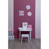A white children's dressing table with a matching stool is positioned against a pink wall. The table features a heart-shaped decoration and is adorned with colorful framed pictures on the wall above. A small storage unit is visible to the left, and the floor is covered with light-colored wood