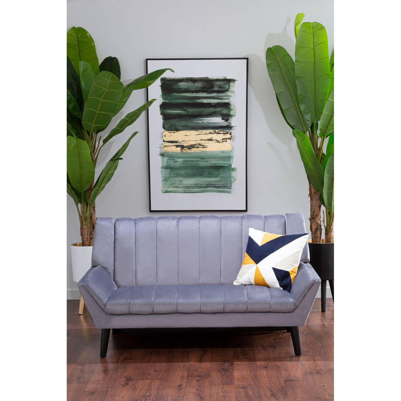 A two-seat grey sofa with a sleek design, featuring a tufted backrest and tapered black legs, is positioned in a modern living space. A decorative pillow with a geometric pattern in yellow, navy, and white is placed on the sofa. Behind the sofa, there is a large framed artwork with abstract green and beige tones, and two tall indoor plants flanking the sides, adding a touch of