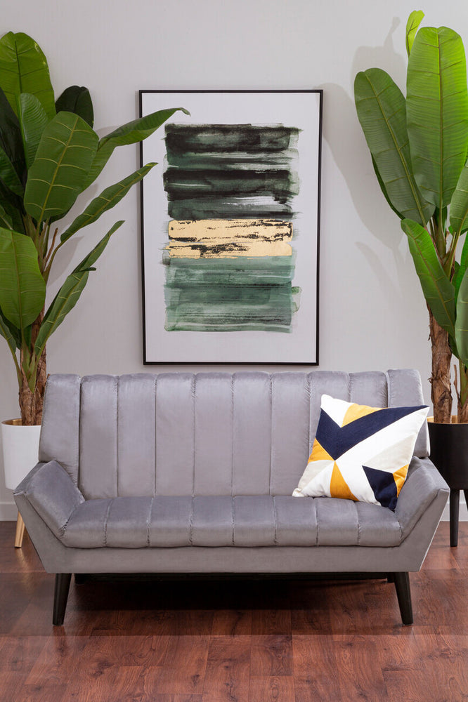 A grey two-seat sofa with a tufted design, positioned in a modern living space. The sofa features black legs and is adorned with a colorful geometric throw pillow. Behind the sofa, there is a large framed abstract artwork and two tall green plants, creating a stylish and inviting atmosphere. The floor is made of warm wood, adding to the cozy ambiance